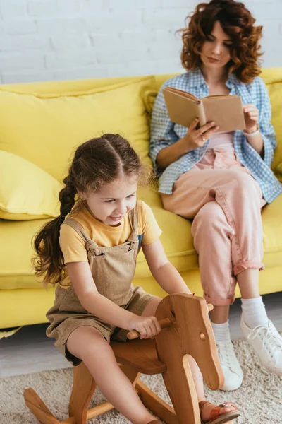 Selective focus of happy kid riding rocking horse while young nanny reading book on yellow sofa — Stock Photo