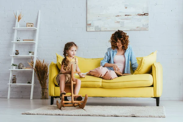 Young babysitter reading book while adorable child riding rocking horse — Stock Photo