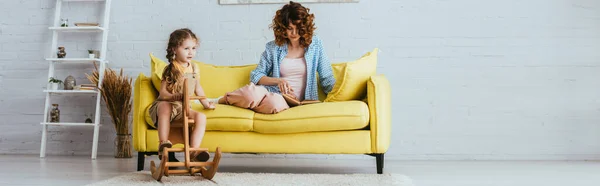 Panoramic shot od adorable child riding rocking horse while nanny reading book on sofa — Stock Photo