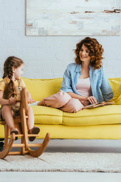 Smiling nanny looking at kid riding rocking horse while sitting on sofa with book — Stock Photo