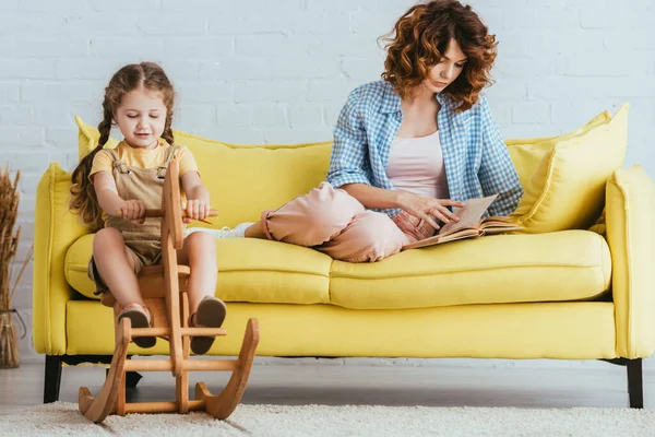 Adorabile bambino cavalcare cavallo a dondolo mentre giovane babysitter lettura libro su divano giallo — Foto stock