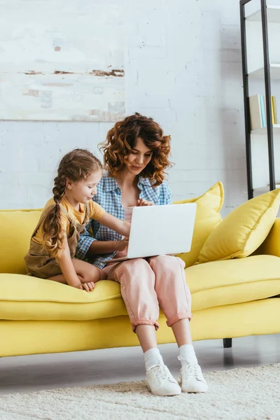 Красивая няня и милый ребенок сидит на желтом диване и с помощью ноутбука — стоковое фото
