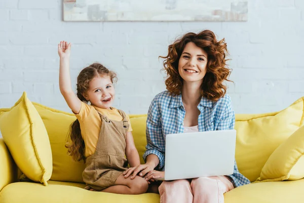 Happy child waving hand and young nanny smiling at camera while sitting on sofa with laptop — Stock Photo