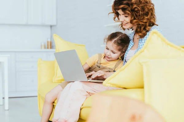 Selective focus of smiling nanny and child sitting on sofa and using laptop — Stock Photo