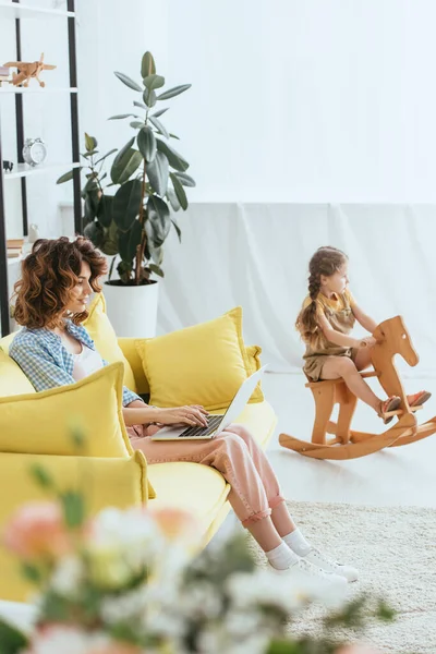 Selektiver Fokus junger Babysitter mit Laptop, während entzückendes Kind Schaukelpferd reitet — Stockfoto