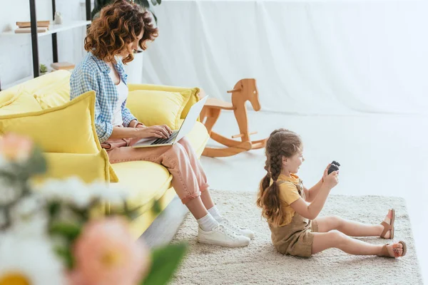 KYIV, UCRANIA - 19 de junio de 2020: enfoque selectivo de la niñera joven que trabaja en el ordenador portátil en el sofá cerca de los niños jugando videojuegos en el suelo - foto de stock