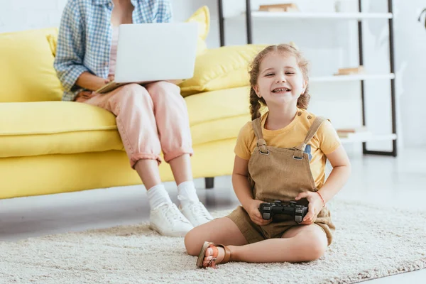 KIEW, UKRAINE - 19. JUNI 2020: Ausschnittansicht eines Kindermädchens, das am Laptop arbeitet, während ein glückliches Kind mit Joystick auf dem Boden sitzt — Stockfoto