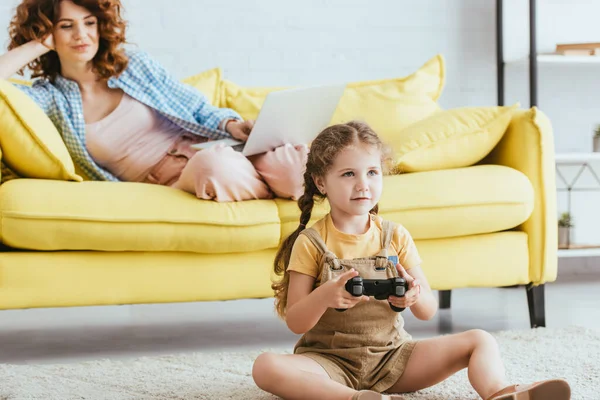 KYIV, UCRANIA - 19 de junio de 2020: enfoque selectivo del niño lindo jugando videojuegos en el suelo cerca de la niñera trabajando en el ordenador portátil en el sofá - foto de stock