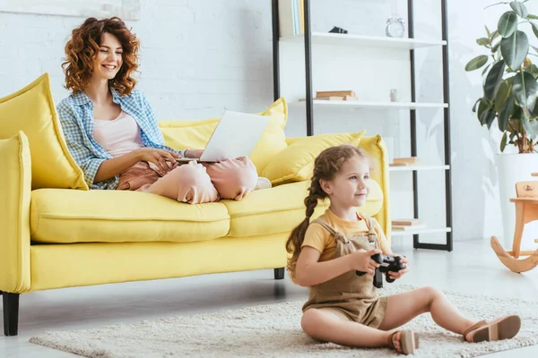 KYIV, UKRAINE - JUNE 19, 2020: smiling nanny using laptop on sofa while happy kid playing video game on floor — Stock Photo