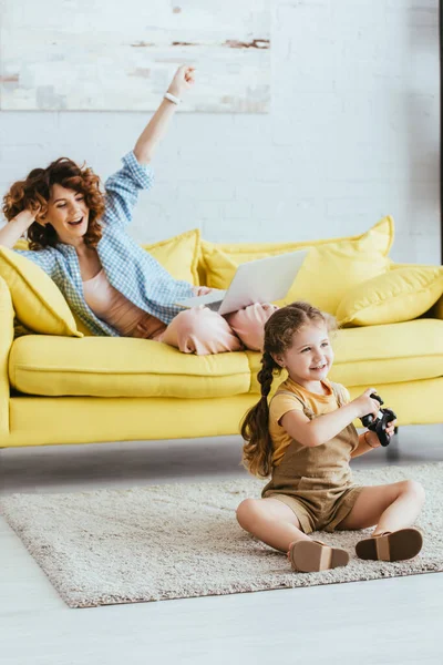 KYIV, UKRAINE - JUNE 19, 2020: selective focus of nanny showing winner gesture while kid sitting on floor with joystick — Stock Photo