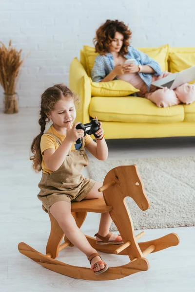 KYIV, UKRAINE - JUNE 19, 2020: selective focus of cute child riding rocking horse with joystick near nanny sitting on sofa with laptop and smartphone — Stock Photo