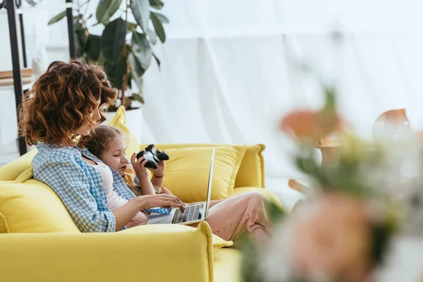 KYIV, UKRAINE - JUNE 19, 2020: selective focus of young babysitter using laptop near kid playing video game — Stock Photo
