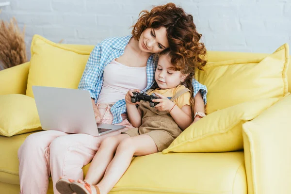 KYIV, UKRAINE - JUNE 19, 2020: young babysitter working on laptop near cute child holding joystick — Stock Photo