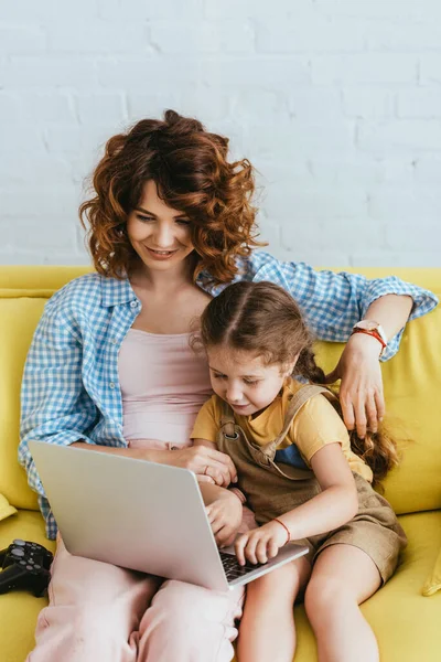 KYIV, UCRANIA - 19 DE JUNIO DE 2020: niñera sonriente y niño usando el ordenador portátil juntos en el sofá cerca de joystick - foto de stock