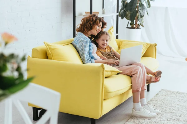 Selective focus of young nanny and cute kid using laptop while sitting on sofa — Stock Photo