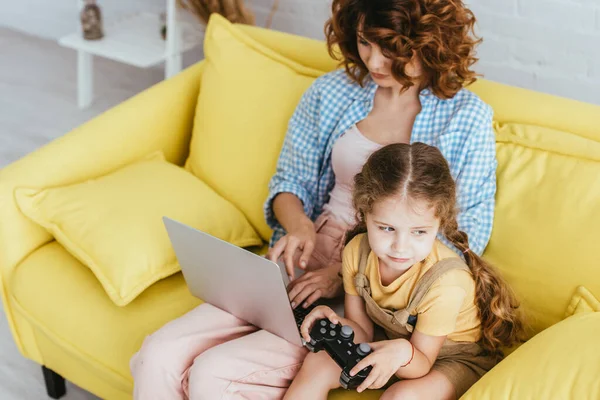 KYIV, UCRAINA - GIUGNO 19, 2020: vista ad alto angolo di bambino carino con joystick vicino alla tata che lavora sul computer portatile — Foto stock