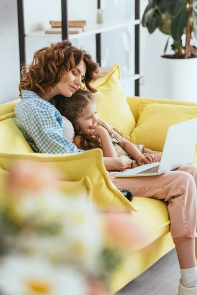 Foyer sélectif de nounou heureuse et adorable enfant en utilisant un ordinateur portable tout en étant assis sur le canapé — Photo de stock