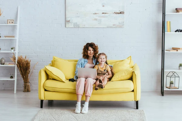 Bonne nounou et adorable enfant assis sur le canapé avec ordinateur portable et souriant à la caméra — Photo de stock