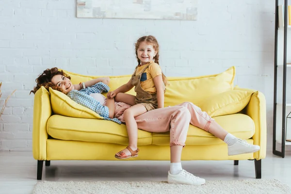 Heureux enfant assis sur couché nounou tout en s'amusant sur canapé et en regardant la caméra — Photo de stock