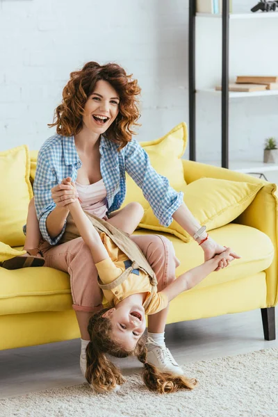 Alegre niñera sosteniendo feliz niño al revés mientras está sentado en amarillo sofá - foto de stock