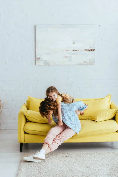 Jovem babá piggybacking feliz criança enquanto sentado no sofá amarelo — Fotografia de Stock
