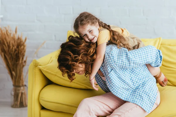 Joven niñera piggybacking alegre niño wihile sentado en amarillo sofá en casa - foto de stock