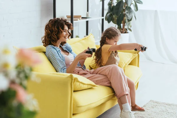 KYIV, UCRANIA - 19 de junio de 2020: enfoque selectivo de la niñera sonriente y el niño jugando videojuegos en el sofá amarillo - foto de stock