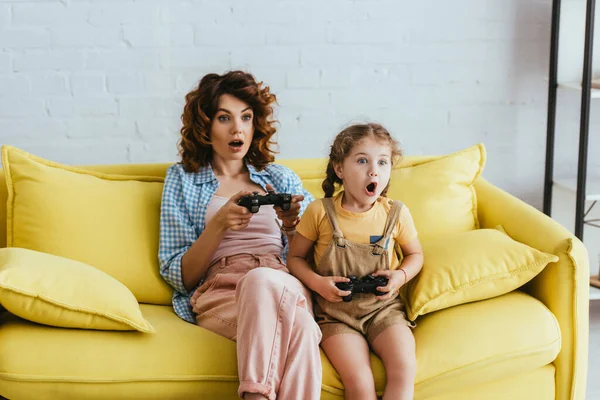 KYIV, UKRAINE - JUNE 19, 2020: excited nanny and adorable kid sitting on yellow sofa with open mouths with joysticks — Stock Photo