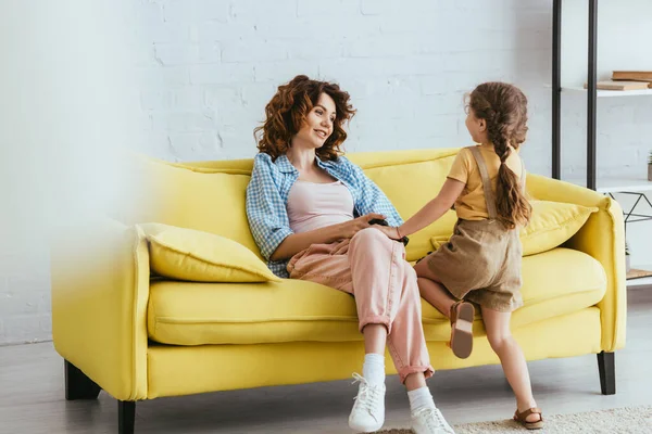 Селективный фокус улыбающейся няни, держащейся за руку с ребенком, сидя на желтом диване — стоковое фото
