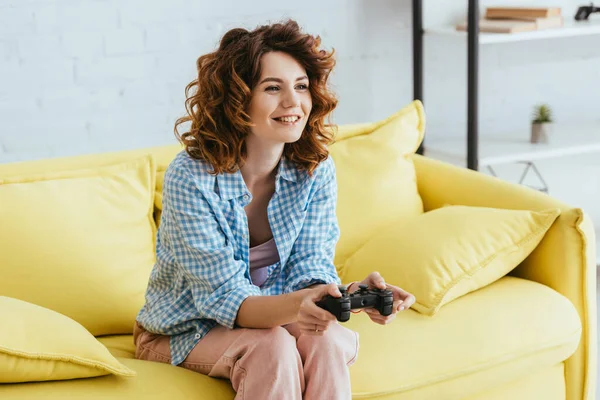 KYIV, UCRANIA - 19 de junio de 2020: mujer atractiva y alegre sentada en el sofá y jugando videojuegos con joystick - foto de stock