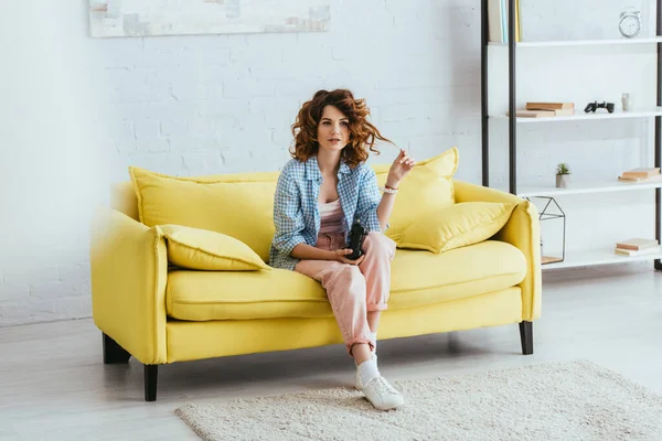 KYIV, UKRAINE - JUNE 19, 2020: selective focus of young woman touching hair while sitting on sofa with joystick — Stock Photo