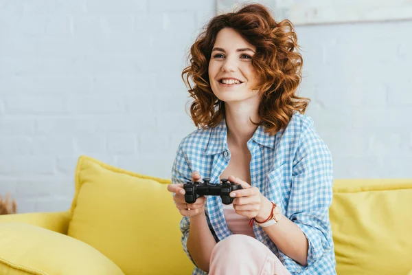 KYIV, UKRAINE - JUNE 19, 2020: smiling young woman sitting on sofa and playing video game with gamepad — Stock Photo