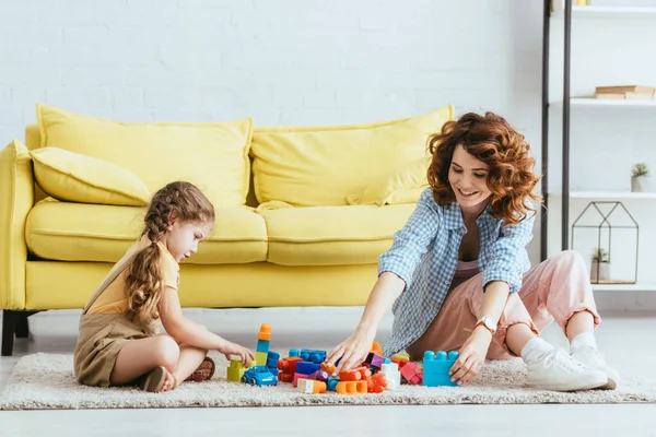 Babysitter sorridente e bambino carino che gioca con i mattoni mentre siede sul pavimento vicino al divano giallo — Foto stock