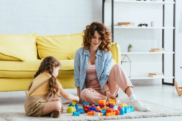 Hermosa niñera y niño sentado en el suelo en la sala de estar y jugando con bloques de construcción - foto de stock