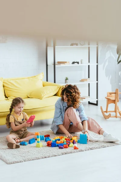 Foco seletivo de criança bonito e jovem babá brincando com blocos de construção enquanto sentado no chão — Fotografia de Stock