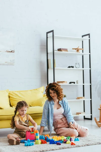 Jeune nounou et adorable enfant assis sur le sol dans le salon et jouer avec les blocs de construction — Photo de stock