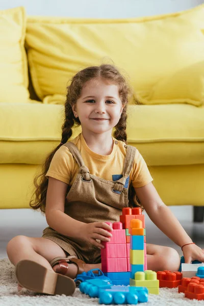 Happy kid smiling at camera while sitting on floor near multicolored building blocks — Stock Photo