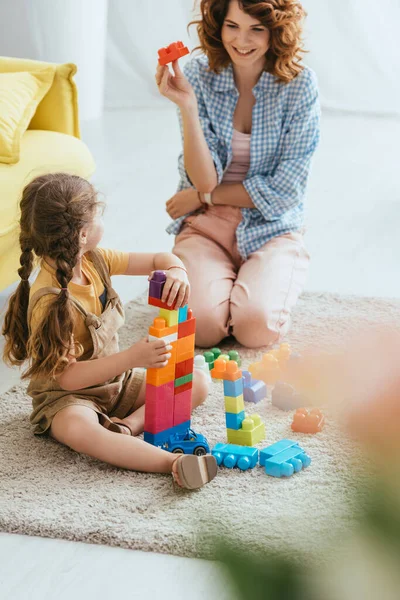 Selektiver Fokus des lächelnden Kindermädchens, das Baustein in der Nähe des Kindes hält, während es gemeinsam auf dem Boden spielt — Stockfoto