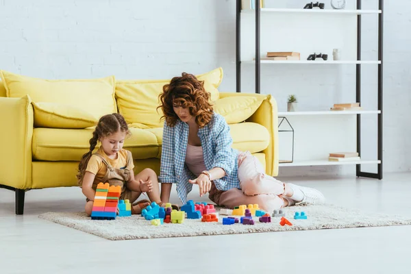 Young nanny and adorable child playing with multicolored building blocks on floor near yellow sofa — Stock Photo