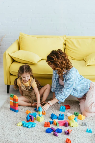 Niñera joven y lindo niño jugando con bloques de construcción multicolores en el suelo cerca de sofá amarillo - foto de stock