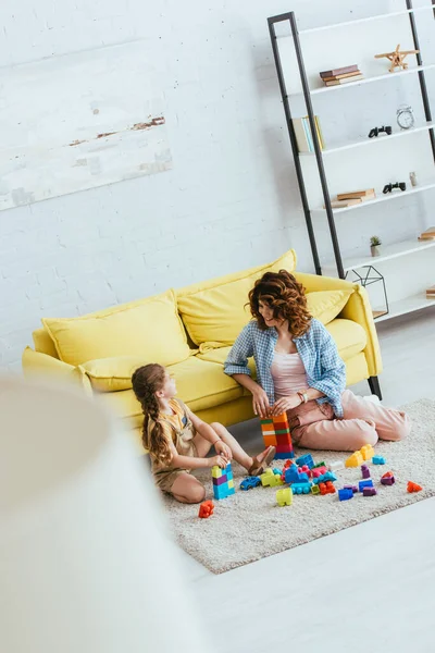 Вибірковий фокус няні і чарівна дитина грає з різнокольоровими будівельними блоками на підлозі у вітальні, високий кут зору — стокове фото