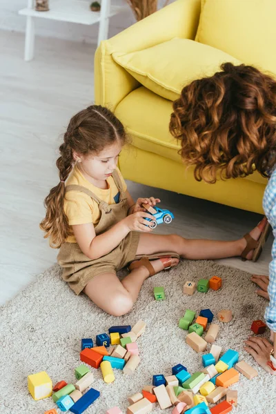 Vista ad alto angolo di carino bambino in possesso di auto giocattolo mentre seduto sul pavimento vicino bambinaia e blocchi multicolore — Foto stock