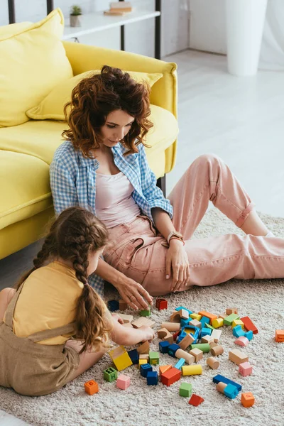 Niñera joven y niño jugando con bloques multicolores en el suelo en la sala de estar - foto de stock