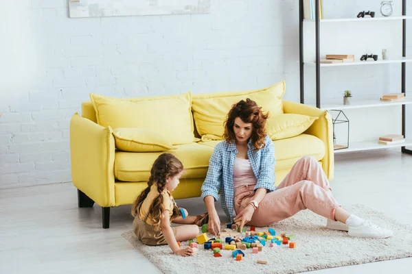 Hermosa niñera y lindo niño jugando con bloques multicolores en el suelo en la sala de estar - foto de stock