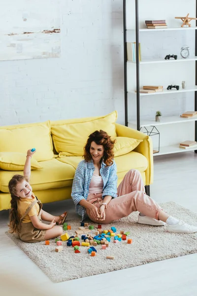 Vue grand angle de l'enfant heureux regardant la caméra tout en étant assis sur le sol près de nounou et cubes multicolores — Photo de stock