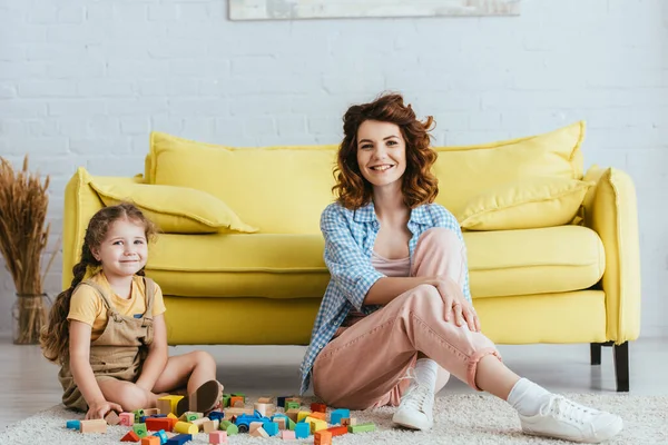 Niñera feliz y lindo niño sonriendo a la cámara cerca de bloques multicolores en el suelo - foto de stock