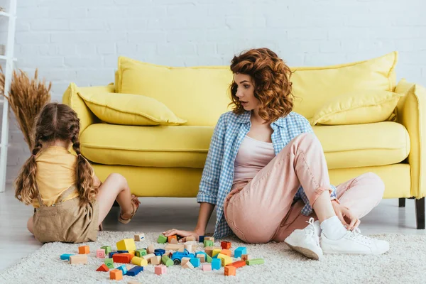 Back view of offended child sitting on floor near young nanny and multicolored blocks — Stock Photo