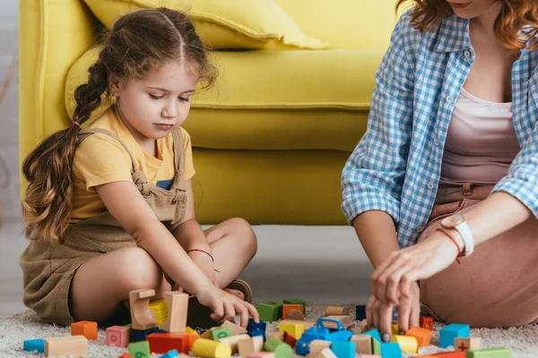 Vista recortada de niñera y adorable niño jugando en el suelo con bloques multicolores - foto de stock