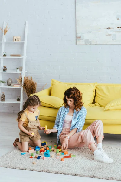 Niñera joven dando bloque a un niño lindo mientras juega en el suelo en la sala de estar - foto de stock