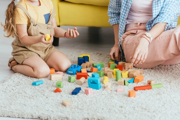 Обрізаний вид на дитину, що тримає різнокольорові блоки біля няні, граючи на підлозі — стокове фото